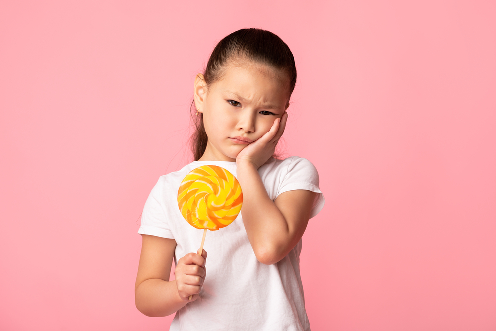 a child holding a lollipop and holding a hand to her face because of pain from a toothache caused by sugar