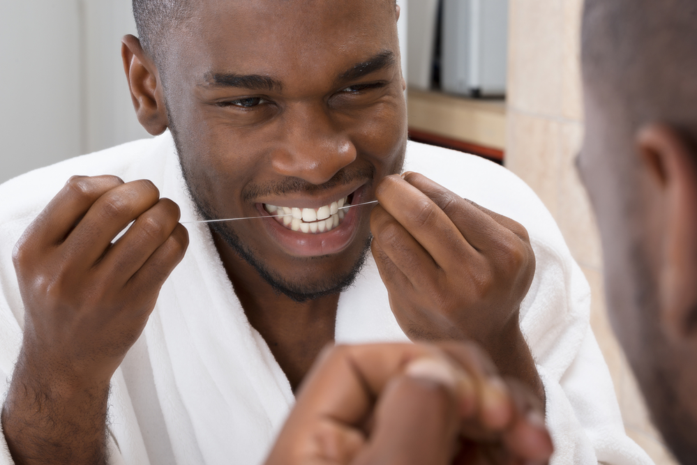 a black man flossing his teeth while looking in the mirror wearing a white bathrobe
