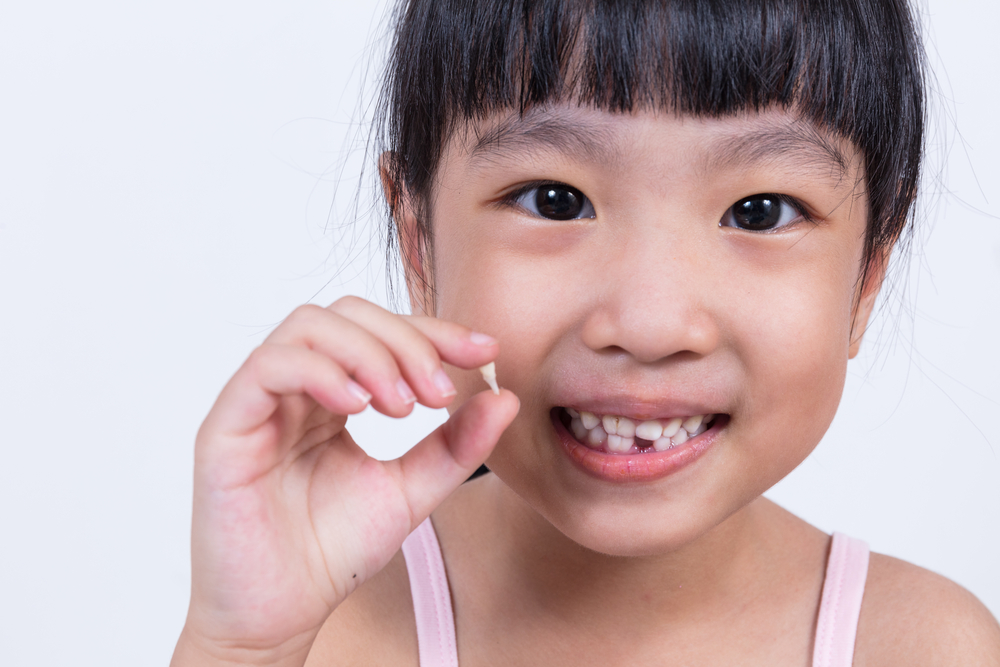 a young Asian girl with a missing bottom tooth holding the tooth that she’s just lost in her hand