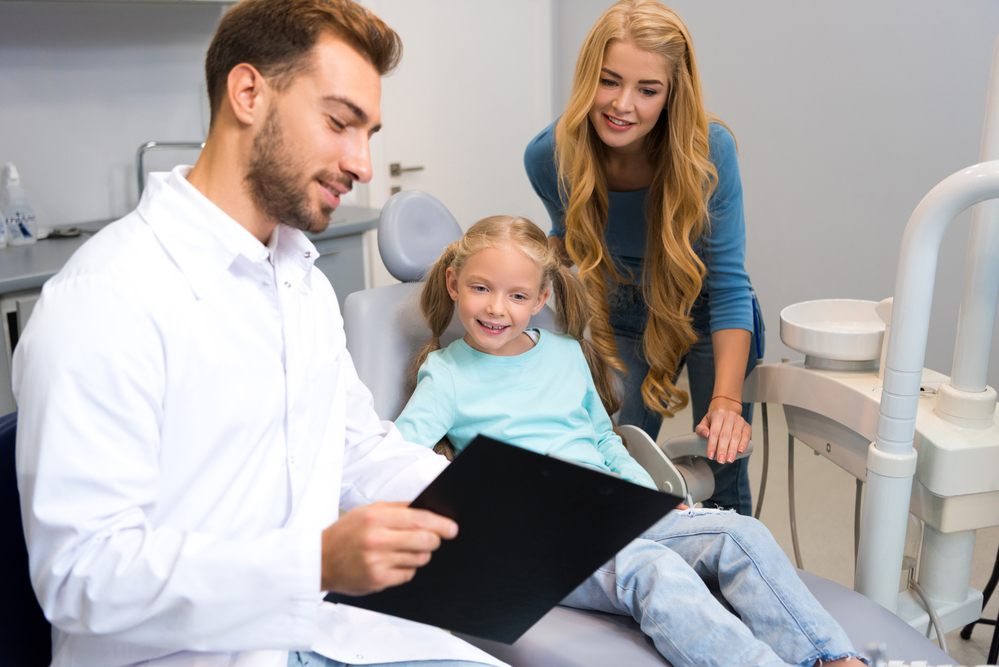 a young girl sitting in a dentist’s chair with the dentist showing her and her mother a chart