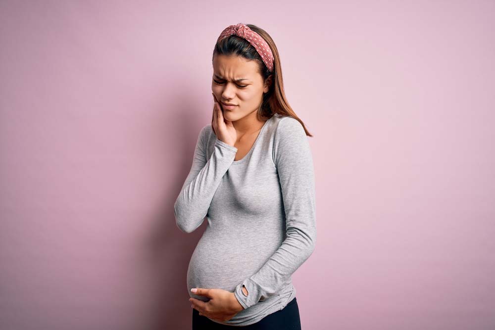 A pregnant woman in a gray shirt stands in front of a pink background, holding her belly with one hand and her face with another, in pain from a toothache.