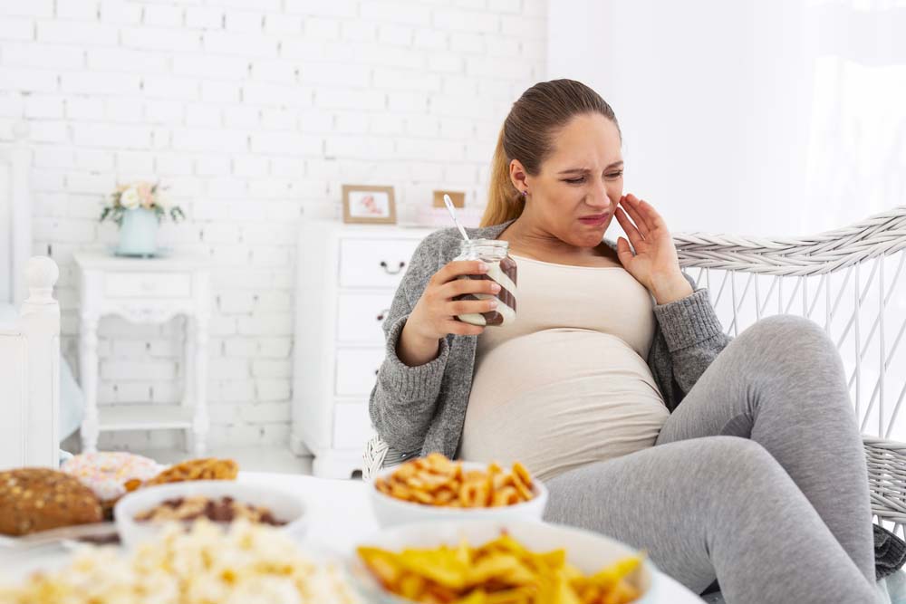 A pregnant woman lies on a couch with a drink in hand, clutching at the side of her face in pain.