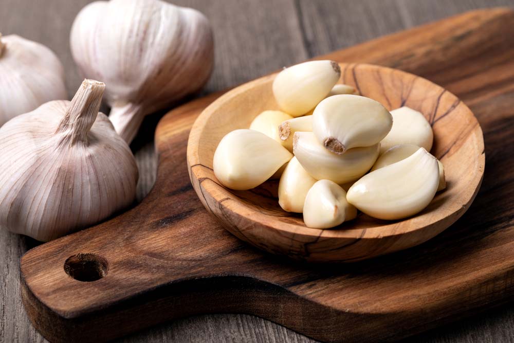 Peeled cloves of garlic in a wooden bowl on top of a wooden cutting board.