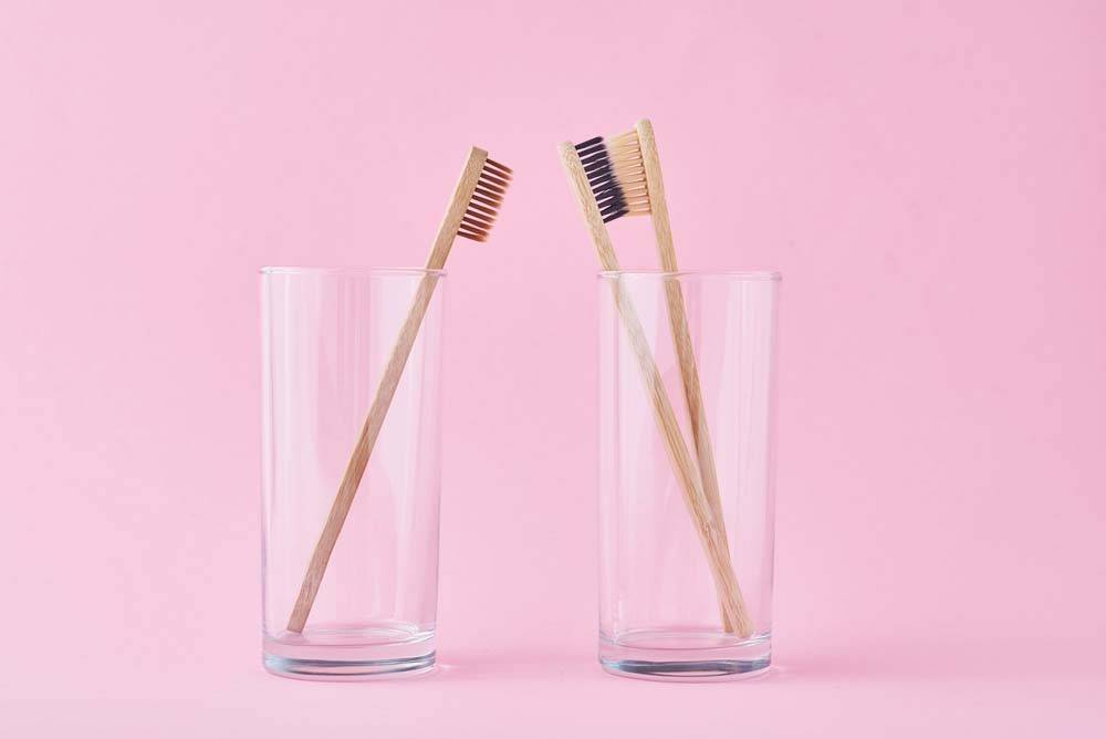 A pink background with two clear cups next to each other with one toothbrush in the cup on the left and two toothbrushes in the right cup.